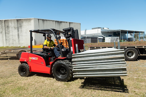 Red ForkForce Enforcer rough terrain forklift lifting steel beams on uneven outdoor site near warehouse, operated by worker in hi-vis gear, highlighting durability for construction.